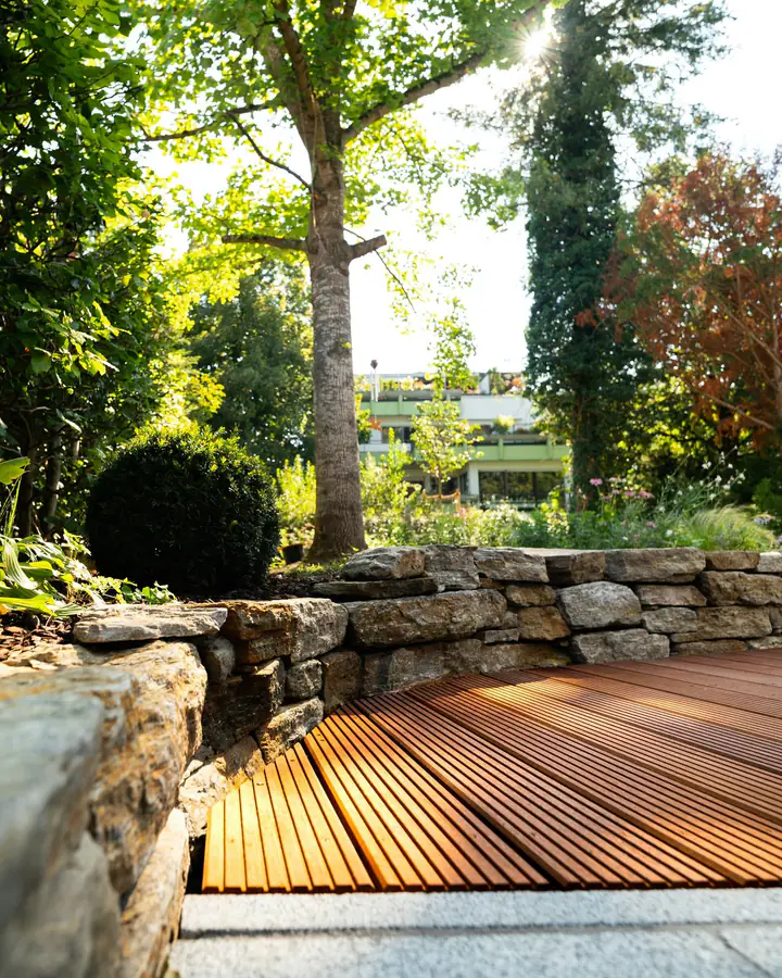 Photo d’illustration : une terrasse en lames de bois posée au pied d’un muret de pierres, dans un jardin arboré.